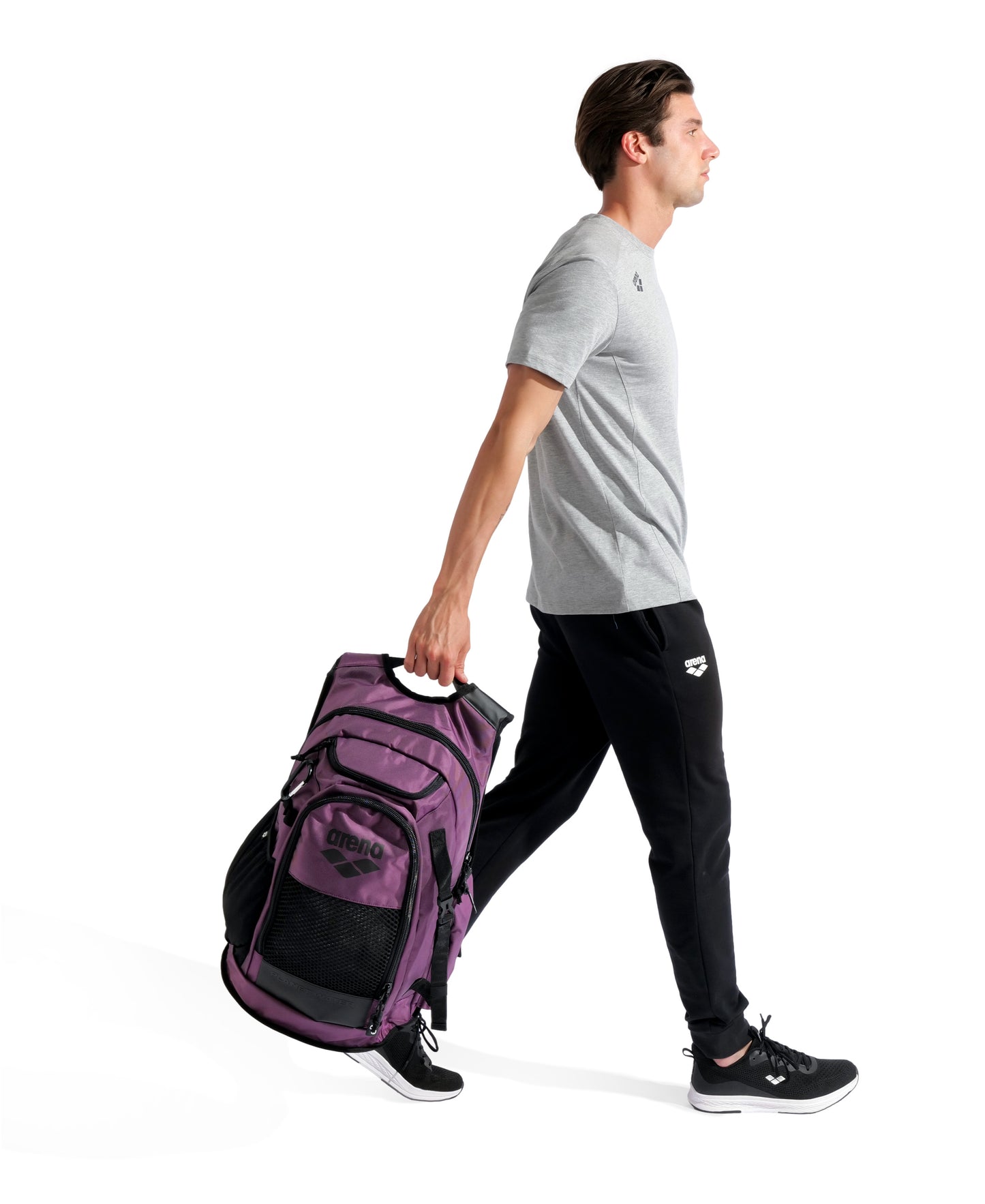 A man in a gray t-shirt, black joggers, and black sneakers walks while pulling the Arena All Set Sports Swim Backpack 45L - Plum by its handle against a plain white background.