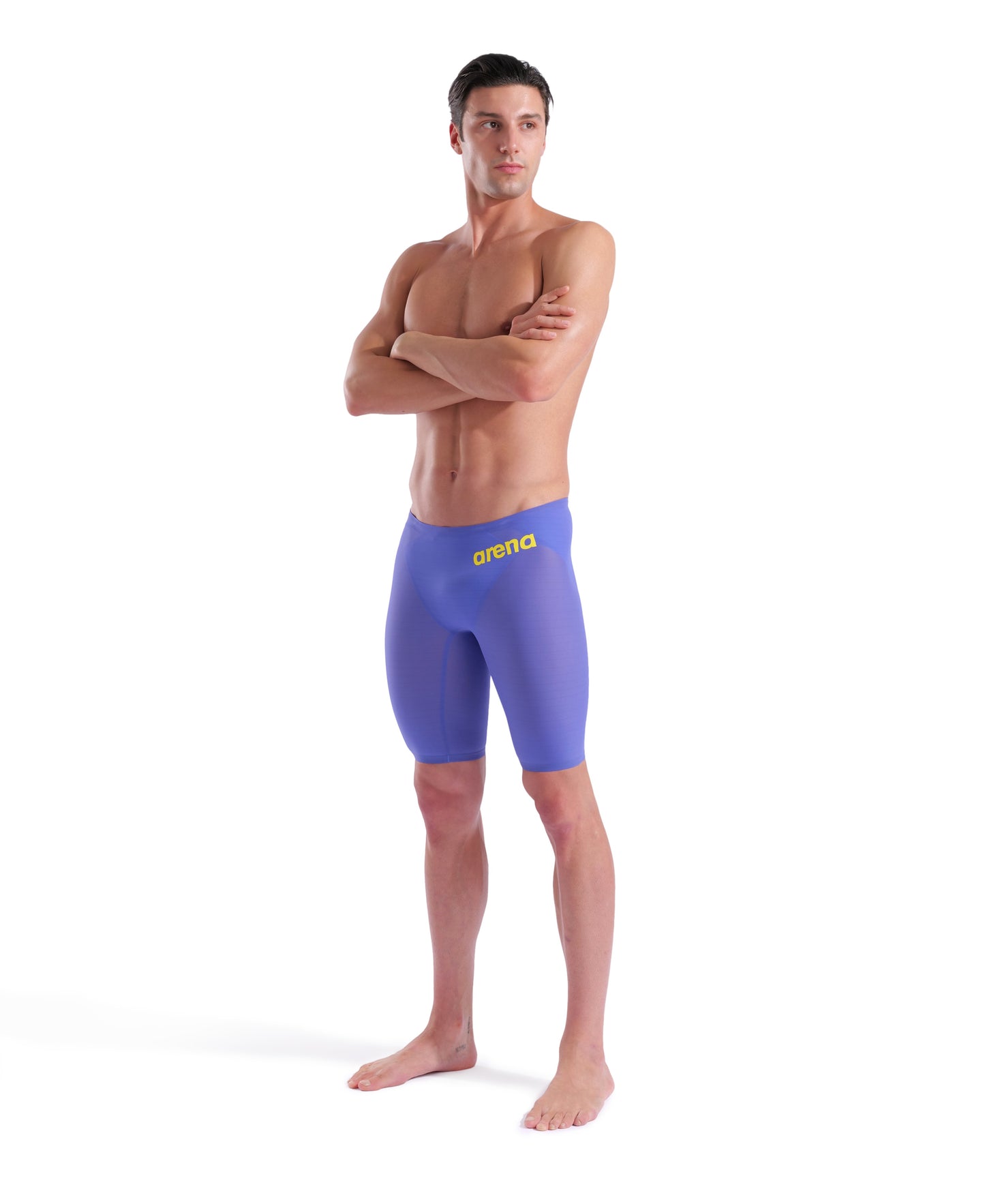 A man stands barefoot with arms crossed, wearing Arena Men's Powerskin Carbon Air2 Jammer – Limited Edition – Eclipse Glare, featuring a yellow "arena" logo, looking off to the side against a plain white background.