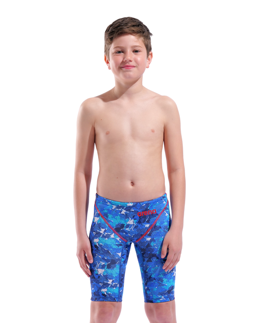 A boy stands smiling, wearing the Arena Boys Powerskin ST Next Jammer - Limited Edition - Splashy by Arena—eco-friendly blue patterned swimwear with red and white accents—made from recycled fabric, against a plain white background.