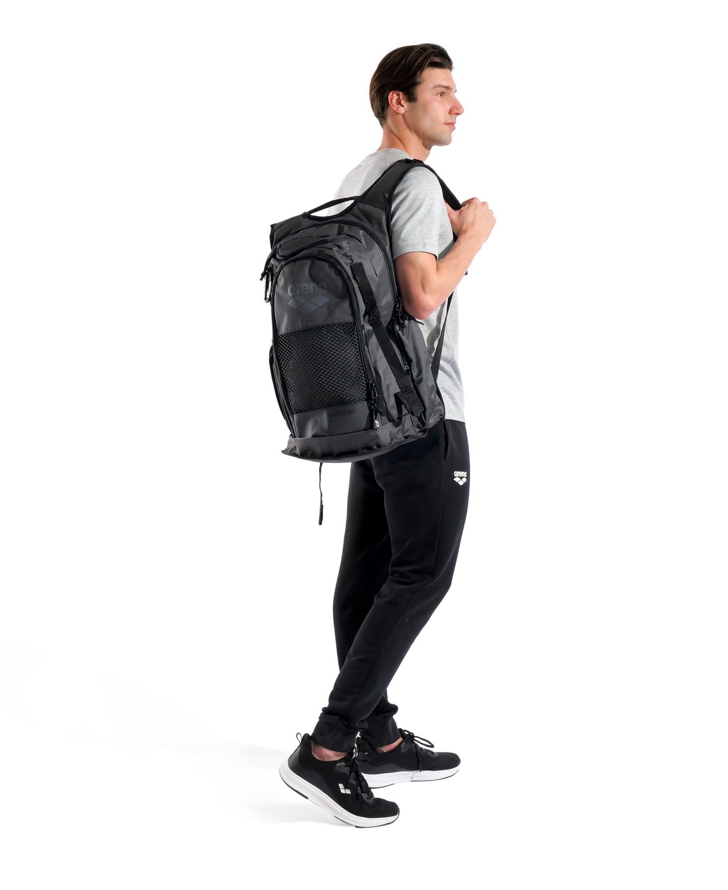 A man in a gray t-shirt, black pants, and black sneakers stands in profile with the Arena All Set Sports Swim Backpack 45L - Black by Arena, featuring a wet management system, slung over one shoulder against a white background.