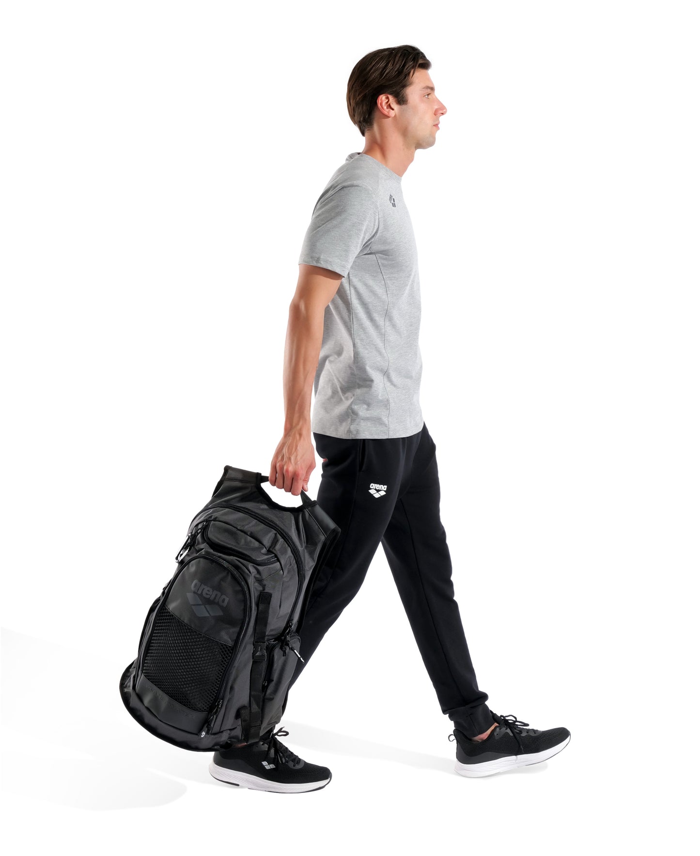 A man in a grey t-shirt and black pants holds the Arena All Set Sports Swim Backpack 45L - Black by Arena by its handle. He wears black sneakers and is photographed against a white background.