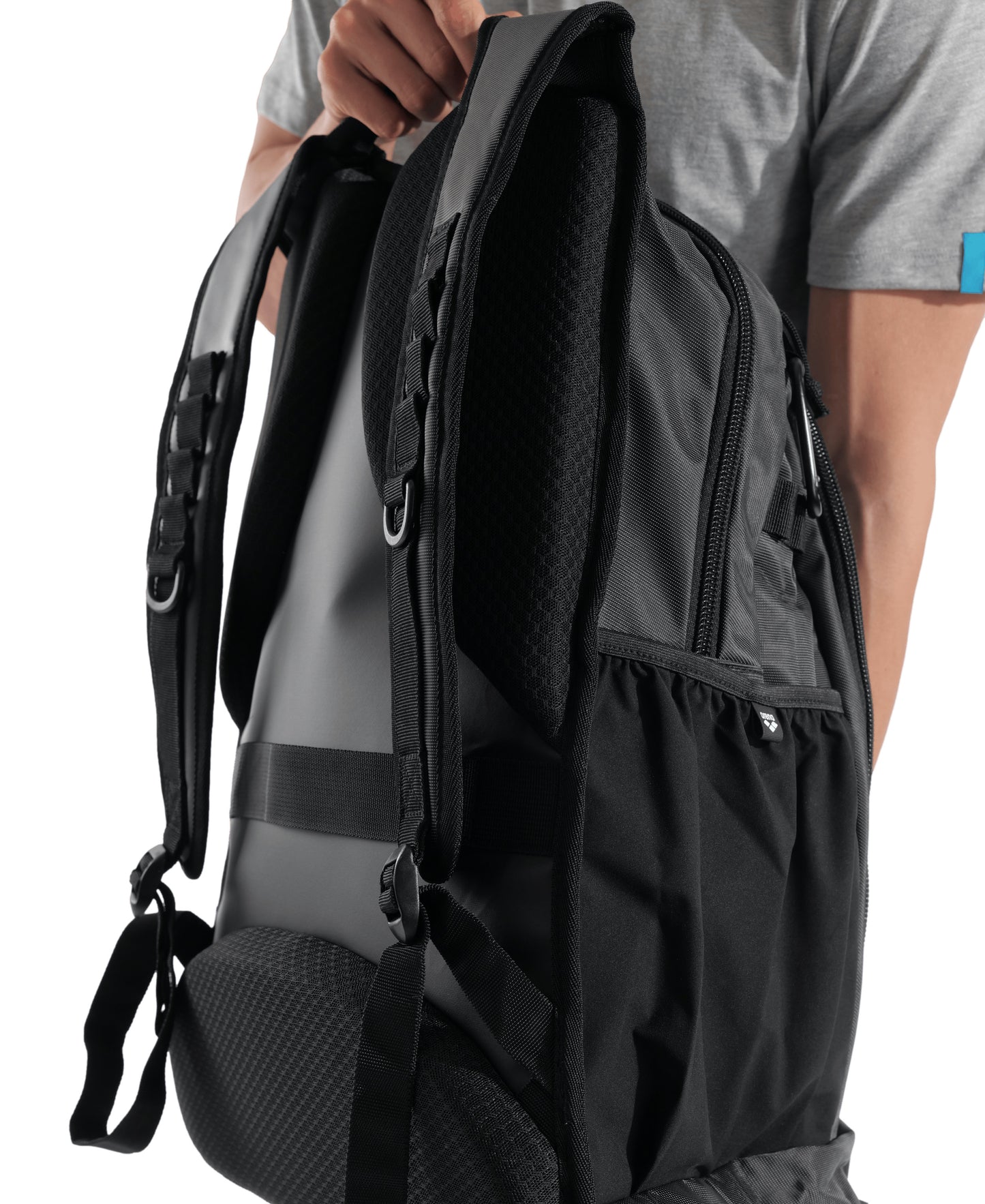 A person in a gray shirt holds the Arena All Set Sports Swim Backpack 45L - Black by one strap, showing its padded back panel, adjustable shoulder straps, and zippered side pocket for wet gear.