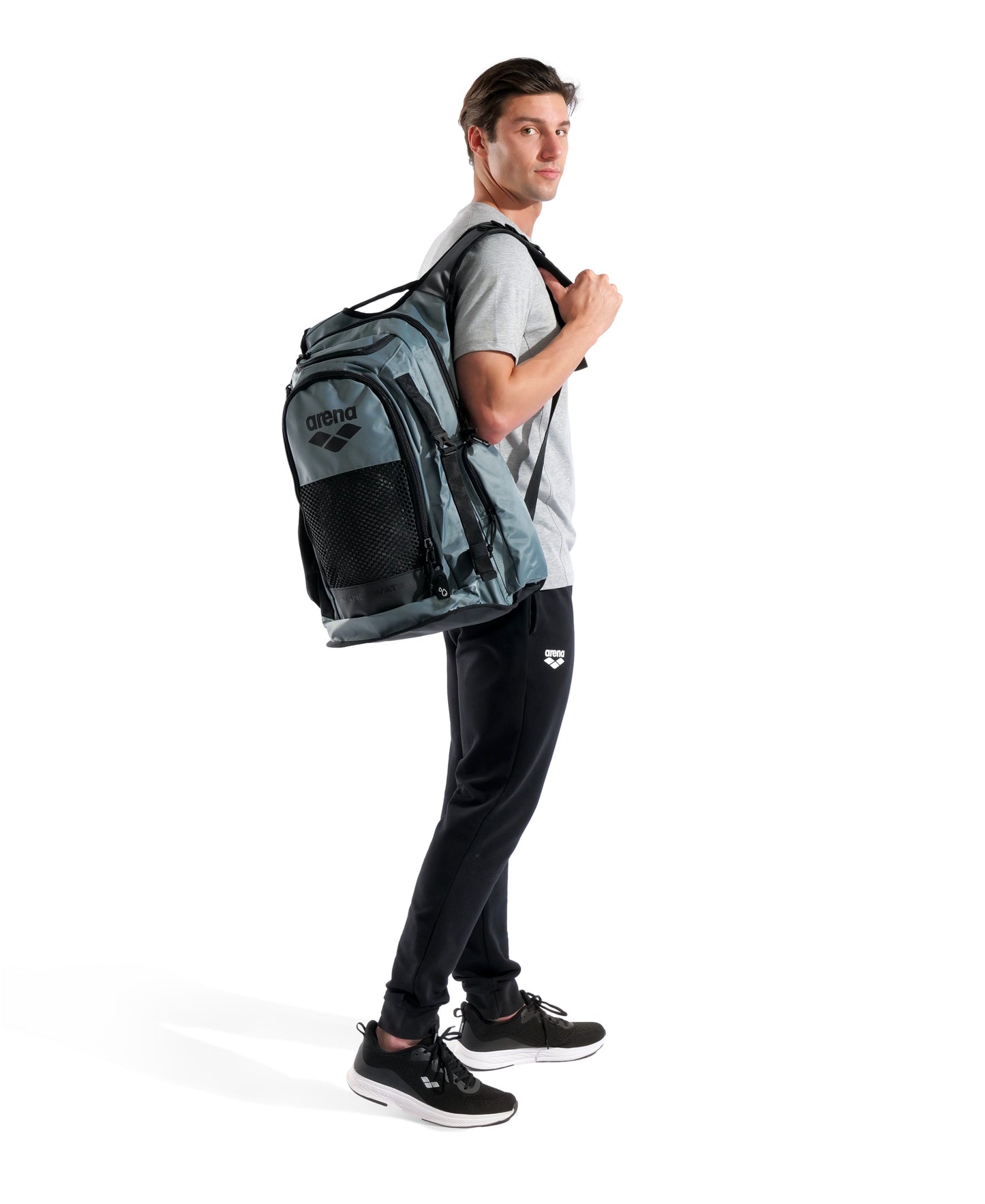 A man in a gray t-shirt, black pants, and black sneakers stands sideways against a white background, carrying the Arena All Set Sports Swim Backpack 45L - Sage with Arena's wet management system on his shoulder.