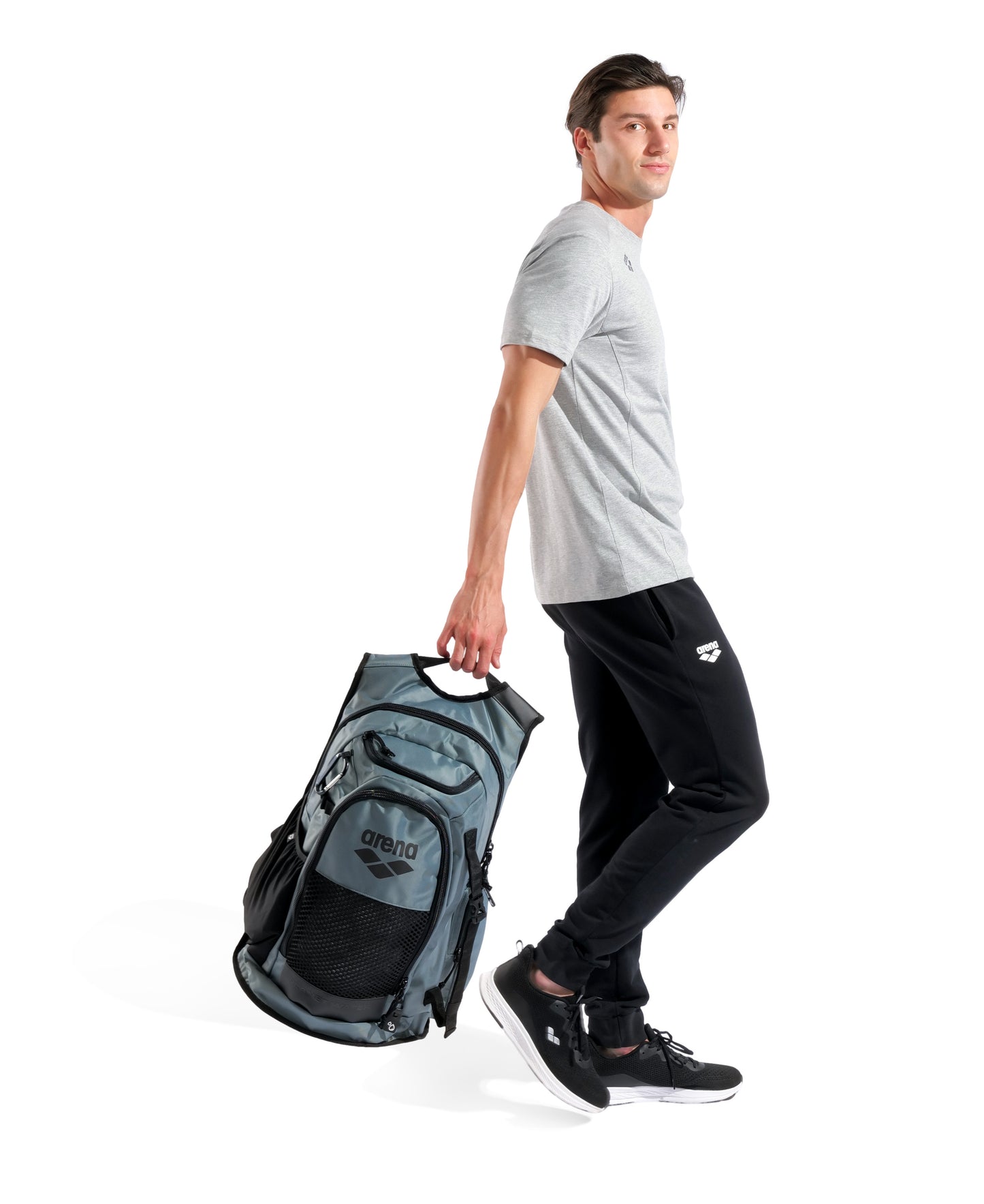 A man wearing a light gray t-shirt, black pants, and sneakers walks holding the Arena All Set Sports Swim Backpack 45L - Sage with wet management system by his side against a white background.