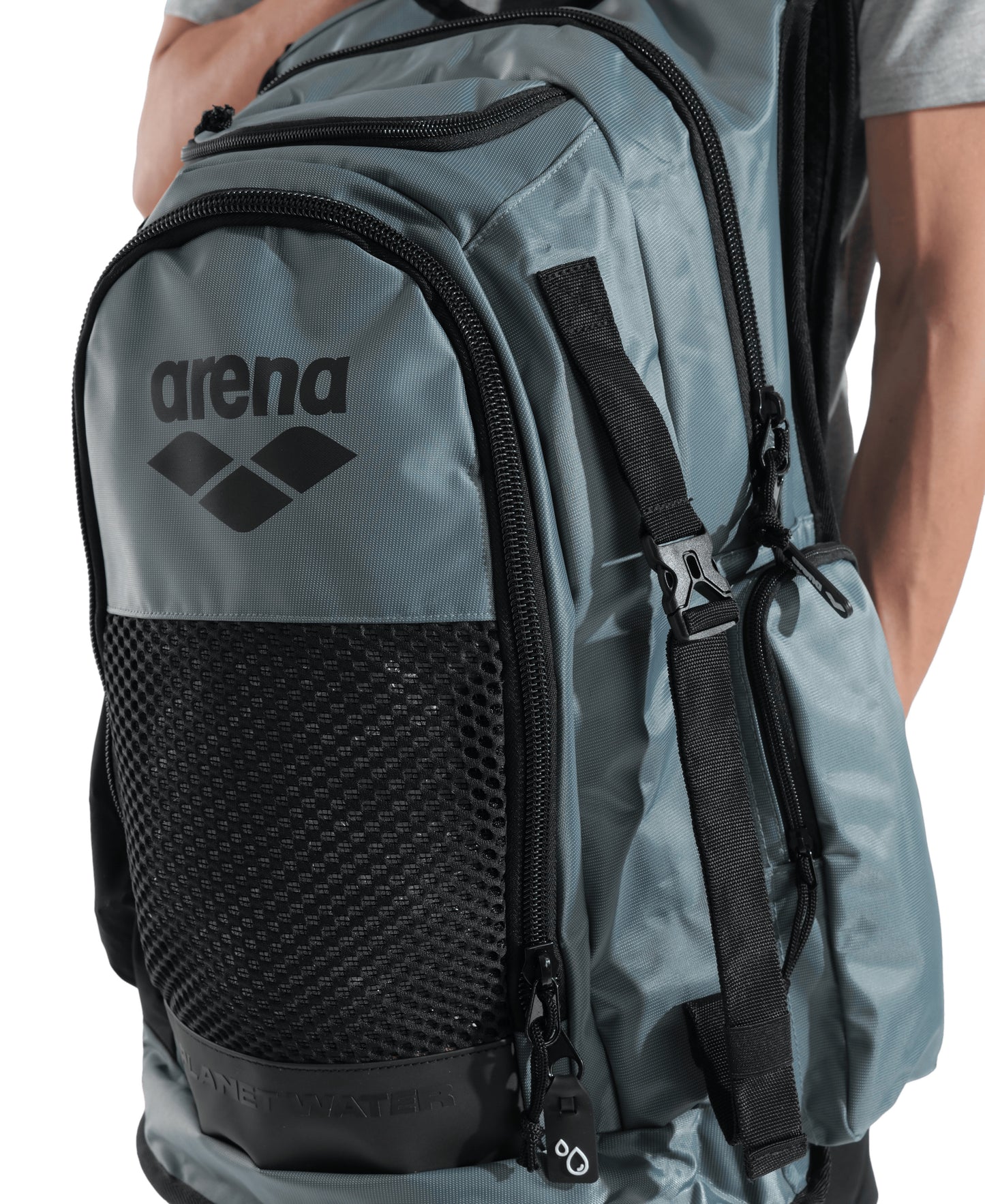 A person wearing a light grey shirt carries the Arena All Set Sports Swim Backpack 45L - Sage, a spacious Arena backpack featuring multiple zippered compartments, a black front mesh pocket, and an integrated wet management system.